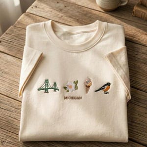 May include: A cream-colored short-sleeve t-shirt featuring embroidered icons of a green bridge, a white flower, a multi-colored ice cream cone, and a bird, with the word MICHIGAN embroidered in brown thread centered below the designs.