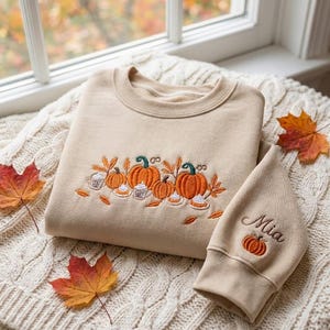 May include: A beige crewneck sweatshirt featuring an embroidered design of orange pumpkins, autumn leaves, and small cups of coffee. The sleeve is embroidered with the name Mia above a small orange pumpkin. The fabric is a soft, solid beige color.