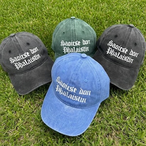 May include: Four baseball caps in shades of blue, green, and gray are arranged on green grass. Each cap features the embroidered text "Saoirse don Phalaistin" in white, with a curved brim and adjustable strap.