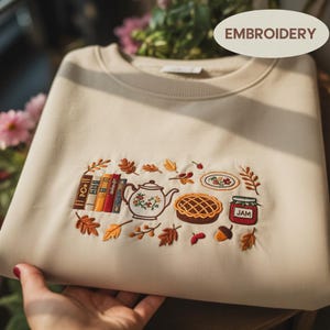 May include: A beige crewneck sweatshirt featuring colorful embroidery of a row of books, a teapot, a pie, a small plate, and a jar labeled JAM, all surrounded by scattered autumn leaves and an acorn, creating a cozy, seasonal aesthetic.