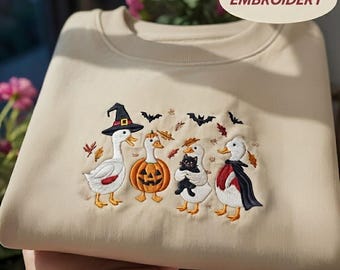 May include: A beige crewneck sweatshirt featuring an embroidered design of four white ducks in Halloween costumes, including a witch hat, a pumpkin suit, a black cat, and a vampire cape, surrounded by small bats and autumn leaves with the text EMBROIDERY.