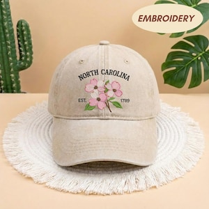 May include: A beige baseball cap with the embroidered words "NORTH CAROLINA" above a floral design. The design includes pink flowers and green leaves. The text "EST. 1789" is below the flowers. The word "EMBROIDERY" is in a circle above the cap.