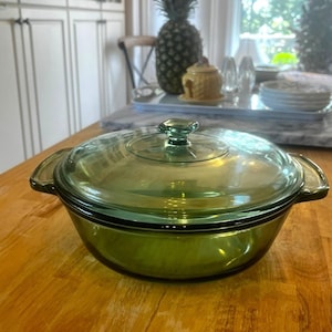 Puede incluir: Una fuente para horno vintage de vidrio verde con tapa de vidrio transparente. El plato tiene dos asas y un pomo cuadrado en la tapa. El plato está sobre una superficie de madera. El tema general es el menaje de cocina.