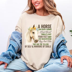 Puede incluir: Camiseta beige de manga larga con una ilustración de un caballo blanco con una corona floral. El texto en la camiseta dice: "A Horse Can Hear a Human Heartbeat From at Least Four Feet Away".