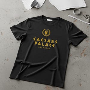 May include: Black crew neck t-shirt with a gold design featuring a laurel wreath, a bust, and the words "CAESARS PALACE LAS VEGAS". The shirt is made of a soft material and has a tag at the neckline.