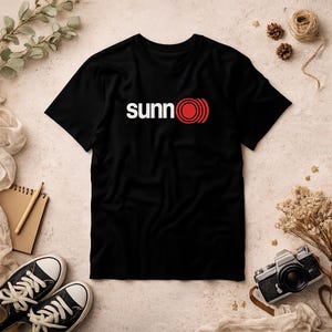 May include: Black t-shirt with the word "sunn" in white, and a red and white circular design. The shirt is made of a soft material and is likely a casual wear item.