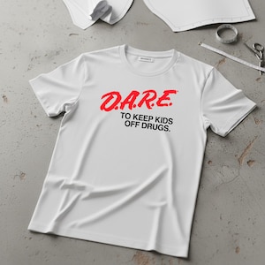 May include: White t-shirt with the red text "D.A.R.E. TO KEEP KIDS OFF DRUGS." The shirt is made of a soft material and has a crew neck. The shirt is laying on a gray surface with sewing tools in the background.