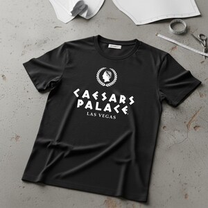 May include: Black crew neck t-shirt with the Caesars Palace Las Vegas logo in white. The logo features a laurel wreath and a bust of a Roman figure. The shirt is made of a soft material and is displayed on a gray surface.
