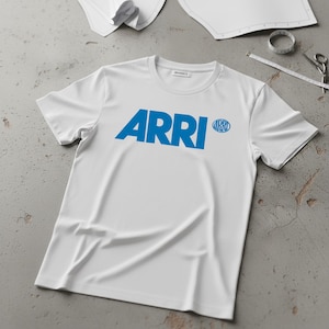 Pode incluir: Camiseta branca de gola redonda com a palavra "ARRI" impressa em letras azuis em negrito no peito. Um logotipo circular menor com "ARRI" está à direita. A camiseta é feita de material macio.