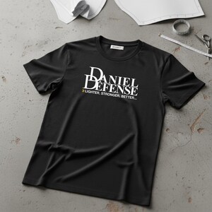 Peut inclure: T-shirt noir à col rond avec le texte blanc "DANIEL DEFENSE" et le slogan "LIGHTER. STRONGER. BETTER...". Le t-shirt est présenté sur une surface grise avec des outils de couture en arrière-plan.