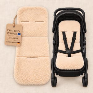 May include: A beige sheepskin pushchair liner with harness slots, measuring 75 x 36 cm, alongside a black pushchair featuring a matching liner. A cardboard tag attached by string reads: HAND-SEWN IN THE EU, 75 x 36 CM, GERMAN BRAND.