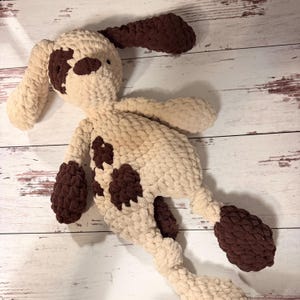 May include: A handmade crochet plush dog toy crafted from thick, soft cream and dark brown yarn. The stuffed animal features long, floppy ears, a cream-colored body with dark brown patches on the face, torso, and paws, and a simple stitched face.