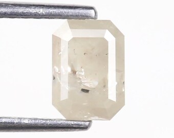 May include: An emerald-cut diamond with a pale, off-white colour and visible internal inclusions. The gemstone is held by a pair of metal tweezers, showcasing its rectangular shape, faceted surface, and translucent, light-toned appearance.