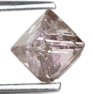 May include: A raw, uncut diamond crystal with a pale pinkish-brown hue and a distinct octahedral shape. The gemstone features natural geometric facets and internal inclusions, held securely between the two metal prongs of a pair of tweezers.