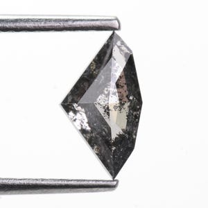 以下が含まれることがあります： ファセット加工が施された凧型のソルト＆ペッパーダイヤモンドのルース。半透明のグレーのボディカラーを持ち、内部全体に黒やダークグレーのインクルージョンが散らばっている。金属製のピンセットで挟まれている。