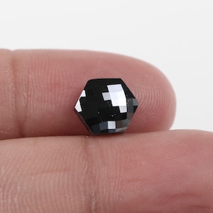 Puede incluir: Una gema negra hexagonal facetada, sostenida entre los dedos. El corte geométrico crea superficies reflectantes. La luz se refleja en las facetas, revelando reflejos y resaltando el color oscuro.