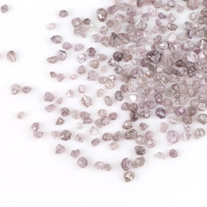 Puede incluir: Una dispersión de pequeños diamantes en bruto, de color púrpura claro, sobre un fondo blanco. Las piedras preciosas varían en tamaño y forma, con un aspecto ligeramente translúcido. Los diamantes están agrupados, creando una superficie texturizada.