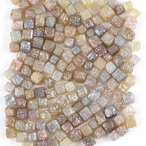 Puede incluir: Una colección de pequeños diamantes de forma irregular en varios colores, incluyendo plata, marrón y amarillo. Los diamantes están dispersos sobre una superficie blanca, creando una apariencia texturizada. Las piedras preciosas son probablemente para la fabricación de joyas.