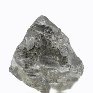 May include: A raw, uncut diamond with a crystalline structure. The stone is a light grey colour and has a textured surface. The diamond is set against a white background.