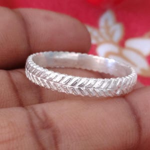Puede incluir: Un anillo de color plateado que presenta un patrón continuo y repetitivo de espiga o chevrón grabado alrededor de toda la banda exterior. La superficie metálica tiene un acabado pulido y reflectante que capta la luz, creando textura.