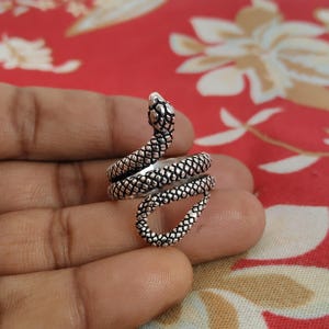 May include: A silver snake ring with a coiled design. The ring features a detailed snake head and body, with textured scales. The ring is shown on a hand against a red floral background.