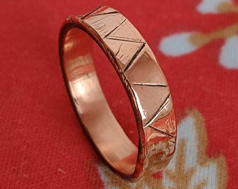 May include: A copper-colored ring with a geometric design. The ring has a rectangular band with etched diagonal lines creating a textured pattern. The ring is set against a red patterned background.
