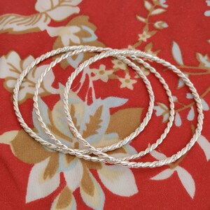 Può includere: Tre braccialetti argentati con design a corda intrecciata. I braccialetti sono circolari e giacciono su un tessuto rosso con un motivo floreale nei toni del bianco, marrone e giallo. I braccialetti sono in metallo lucido.