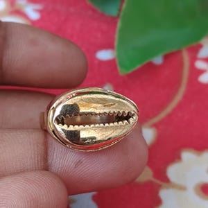 Puede incluir: Un anillo de tono dorado con un diseño de concha de cauri. La concha tiene un acabado metálico liso con una abertura horizontal central que muestra un borde dentado similar a dientes a lo largo del medio, característico de la concha.