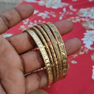 May include: A set of six gold-colored bangles, each with a unique embossed floral or geometric pattern. The bangles are displayed against a red background with a floral design. The bangles are made of metal and are likely worn as jewelry.