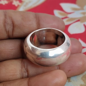 May include: A wide, polished silver-toned band ring with a smooth, rounded exterior surface. The interior of the ring features a small, stamped hallmark visible on the inner wall, indicating the metal purity of the piece in a simple, clean design.