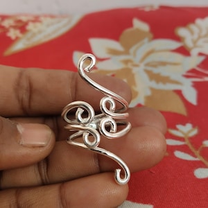 Può includere: Un anello color argento con un design a spirale e aperto. L'anello presenta più anelli e curve, creando un aspetto astratto e artistico. Il metallo ha una finitura lucida, che riflette la luce. Il design dell'anello ricorda una forma floreale o organica stilizzata.