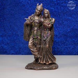 May include: A bronze-toned statue depicting two figures standing together. The male figure wears a crown and holds a flute, while the female figure stands beside him. Both are adorned with intricate garlands and detailed clothing textures.