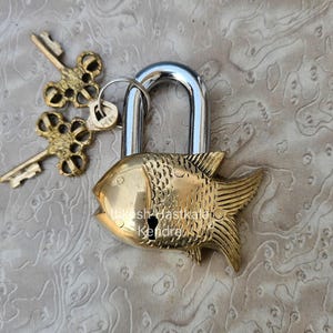 May include: A decorative padlock with a fish-shaped brass body and a silver shackle. Two ornate brass keys are included. The padlock has the text "Ukesh Hastkala Kendra" engraved on it.