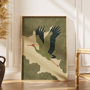 May include: A framed art print featuring a crane in flight. The bird has olive green feathers and a red beak, set against a cream background with stylized cloud patterns. The frame is a light gold color.
