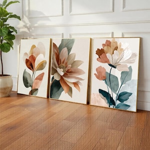 May include: Three framed floral art prints. Each print features a watercolour-style flower in shades of peach, brown, and white, with green and blue-green leaves. The frames have a gold finish, and the artwork is displayed on a light background.