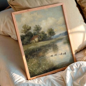 May include: A framed landscape painting featuring a serene river scene with a small house, trees, and ducks. The artwork is in a wooden frame and rests on a bed with pillows.