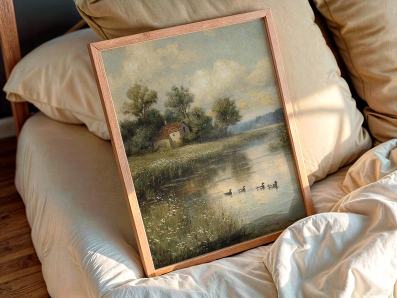 May include: A framed landscape painting featuring a serene river scene with a small house, trees, and ducks. The artwork is in a wooden frame and rests on a bed with pillows.
