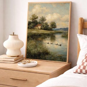 May include: A framed landscape painting depicts a serene river scene with a small house, trees, and ducks. The artwork is displayed on a wooden nightstand, alongside a decorative lamp, books, and a small tray with gold-rimmed glasses. The overall aesthetic is warm and inviting.