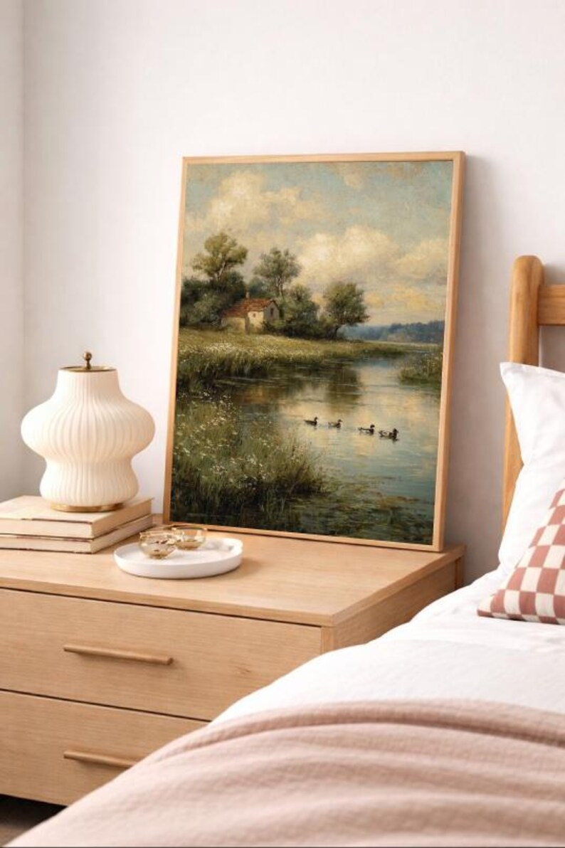 May include: A framed landscape painting depicts a serene river scene with a small house, trees, and ducks. The artwork is displayed on a wooden nightstand, alongside a decorative lamp, books, and a small tray with gold-rimmed glasses. The overall aesthetic is warm and inviting.