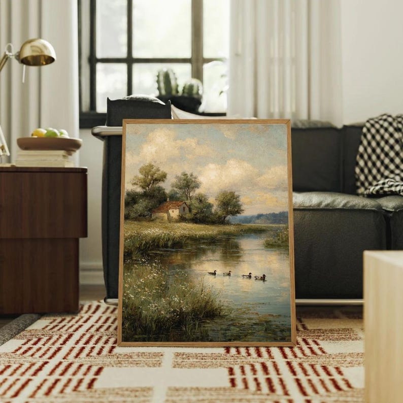 May include: A framed landscape painting depicts a serene river scene with a small house, trees, and ducks. The artwork features a wooden frame and is displayed on a patterned rug, with a sofa and window in the background.