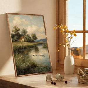 May include: A framed landscape painting depicts a serene river scene with a small house, trees, and ducks. The artwork is in a wooden frame and leans against a wall. A white vase with yellow branches sits near a window.