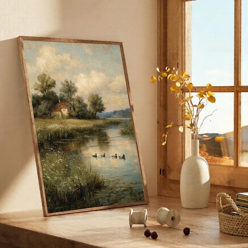 May include: A framed landscape painting depicts a serene river scene with a small house, trees, and ducks. The artwork is in a wooden frame and leans against a wall. A white vase with yellow branches sits near a window.