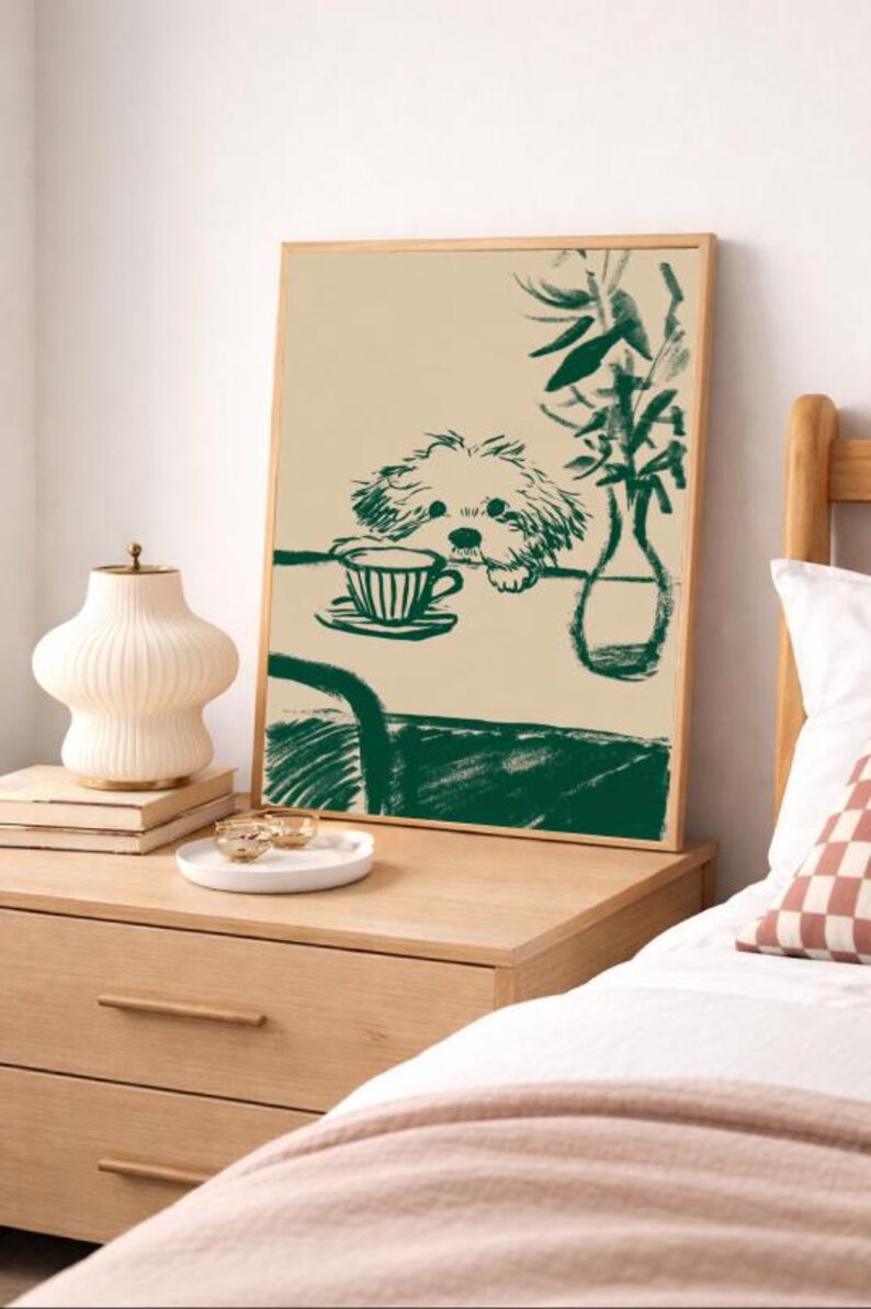 May include: A framed art print featuring a green sketch of a dog peering over a table with a teacup. The artwork is in a wooden frame and is displayed on a light-colored wall. A bedside table with a lamp and decorative items is in the foreground.