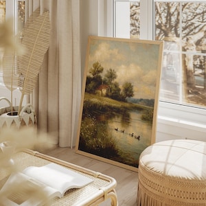 May include: A framed landscape painting depicts a serene waterside scene with a small house, trees, and ducks. The artwork is displayed in a light-colored wooden frame, leaning against a wall near a window. The room also features a woven basket, a bench, and a round ottoman.