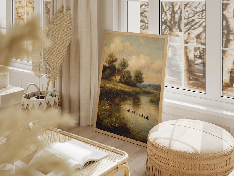 May include: A framed landscape painting depicts a serene waterside scene with a small house, trees, and ducks. The artwork is displayed in a light-colored wooden frame, leaning against a wall near a window. The room also features a woven basket, a bench, and a round ottoman.