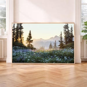 May include: A framed landscape print featuring a mountain scene with evergreen trees and a field of wildflowers. The artwork is displayed on a wooden floor, with potted plants and windows in the background.