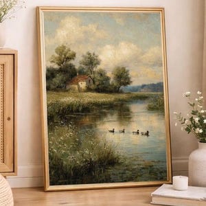 May include: A framed landscape painting depicts a serene waterscape with a small house nestled among trees. Ducks swim in the water, reflecting the sky's soft blues and whites. The gold frame adds a classic touch to the artwork.