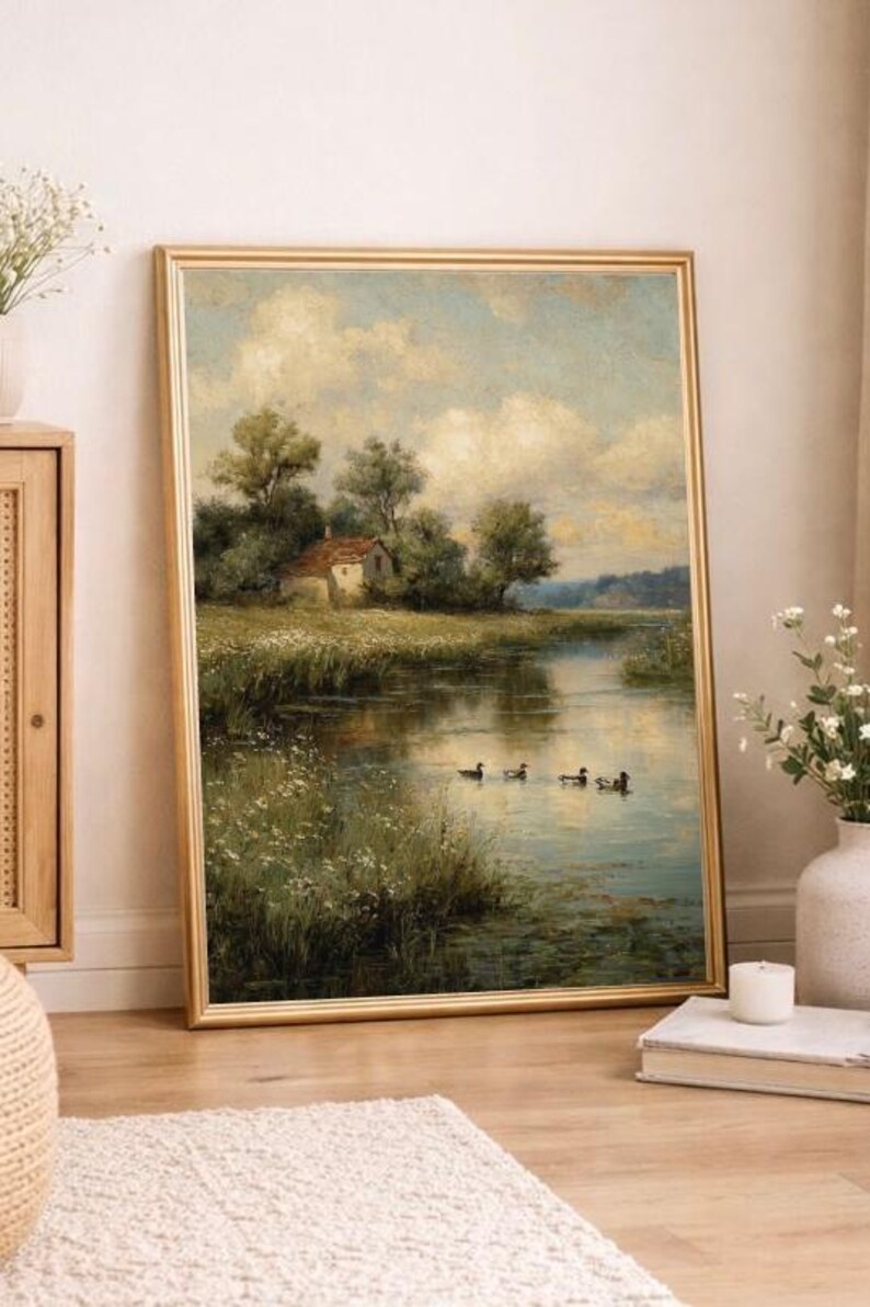 May include: A framed landscape painting depicts a serene waterscape with a small house nestled among trees. Ducks swim in the water, reflecting the sky's soft blues and whites. The gold frame adds a classic touch to the artwork.