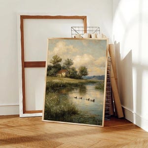 May include: A framed landscape painting depicts a serene waterscape with a small house nestled among trees. Ducks swim in the water, reflecting the sky. The artwork is in a gold frame, leaning against a wall with other art supplies.