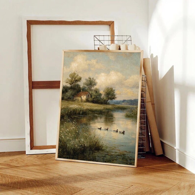 May include: A framed landscape painting depicts a serene waterscape with a small house nestled among trees. Ducks swim in the water, reflecting the sky. The artwork is in a gold frame, leaning against a wall with other art supplies.